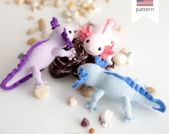 Op de afbeelding: Drie gehaakte axolotl knuffels in verschillende kleuren: paars, roze en blauw. De knuffels liggen op een wit oppervlak met kleine stenen en schelpen.