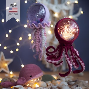 May include: Crocheted jellyfish, octopus, and whale ornaments with clear glass globes. The jellyfish is lavender and white, the octopus is burgundy and white, and the whale is pink. The ornaments are lit with fairy lights. The image includes the text "PDF pattern".