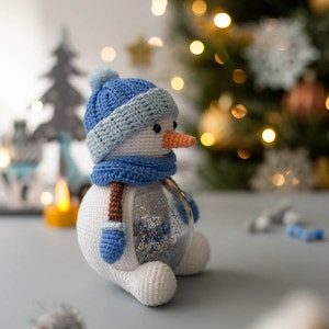 May include: A handmade snowman ornament with a blue hat, scarf, and mittens. The snowman has a carrot nose and a clear glass body filled with glitter. The ornament is crocheted and has brown suspenders. A festive holiday decoration.