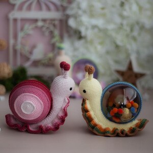 May include: Two crocheted snail toys. One snail is pink and white with a spiral shell. The other snail is yellow with a clear plastic shell filled with colorful pom-poms.