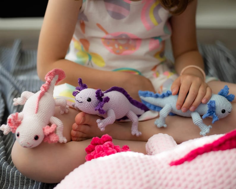 K&ouml;nnte beinhalten: Ein Kind h&auml;lt drei geh&auml;kelte Axolotl-Pl&uuml;schtiere. Die Axolotl sind rosa, lila und blau. Das Kind tr&auml;gt ein wei&szlig;es Kleid mit einem bunten Muster.