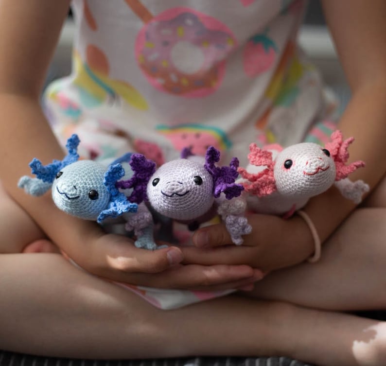 K&ouml;nnte beinhalten: Drei geh&auml;kelte Axolotl-Pl&uuml;schtiere in verschiedenen Farben: hellblau, lavendel und rosa. Die Pl&uuml;schtiere haben gro&szlig;e, runde Augen und kleine, l&auml;chelnde M&uuml;nder.