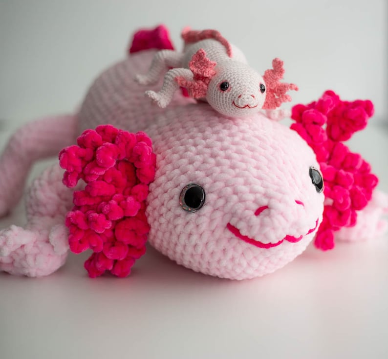 K&ouml;nnte beinhalten: Ein rosa geh&auml;keltes Axolotl-Pl&uuml;schtier mit einem kleineren rosa geh&auml;kelten Axolotl-Pl&uuml;schtier auf dem Kopf.