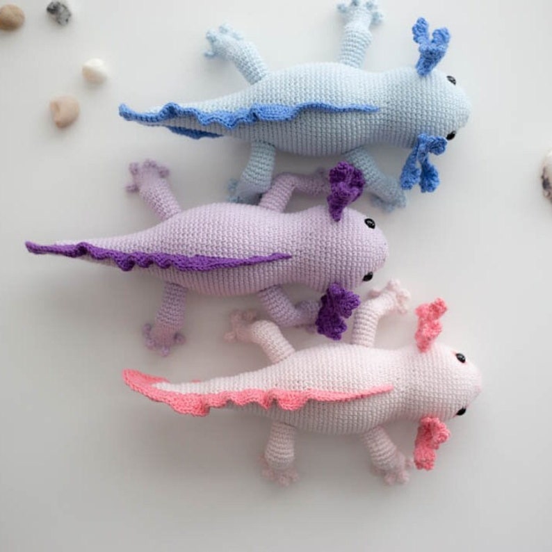 K&ouml;nnte beinhalten: Drei geh&auml;kelte Axolotl-Pl&uuml;schtiere in verschiedenen Farben: hellblau, Lavendel und rosa. Jedes Axolotl hat einen gekr&auml;uselten Schwanz und gefiederte Kiemen.