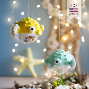 Puede incluir: Dos adornos de pez globo tejidos a ganchillo, uno amarillo y otro verde menta, con vientre blanco, púas texturizadas y grandes ojos circulares. Los peces están unidos a bases esféricas transparentes y cuelgan de hilos finos. Incluye etiqueta de patrón PDF.
