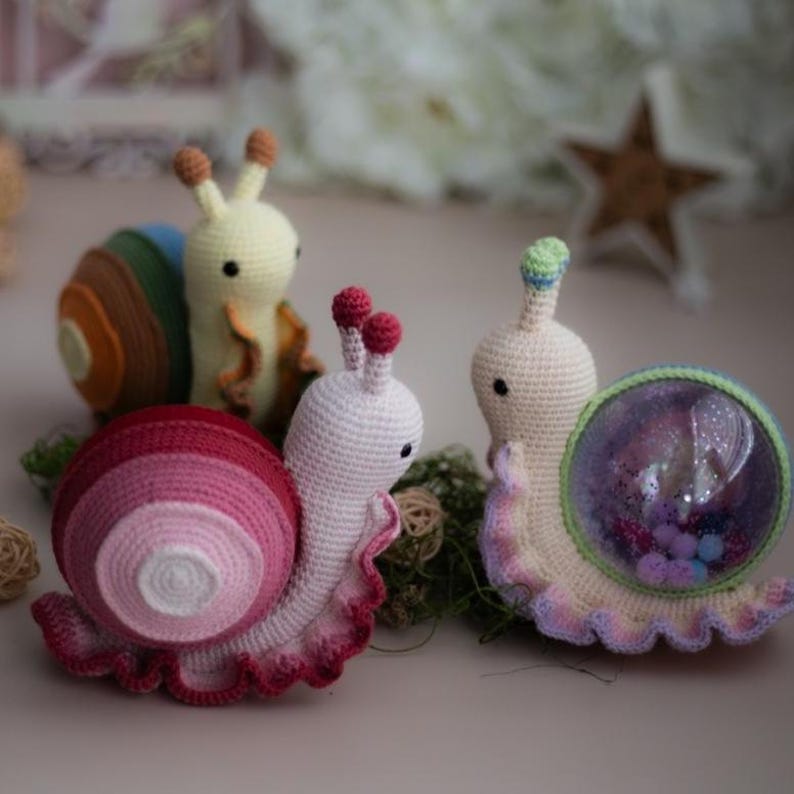 May include: Three crocheted snail toys. One is pink and white with a ruffled shell, one is cream with a green and brown shell, and one is cream with a clear plastic shell filled with glitter and colorful beads.