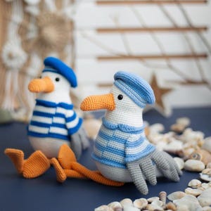 May include: Two handmade crochet seagull toys. Each seagull has a white body, blue and white striped shirt, blue beret, orange beak and feet, and grey wings. The toys are sitting on a blue surface with seashells.