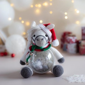 May include: A crocheted white horse ornament with a Santa hat, a green and red scarf, and a clear glass body filled with white snow-like material. The horse has gray hooves and a gray muzzle. Two small silver bells hang from the scarf.