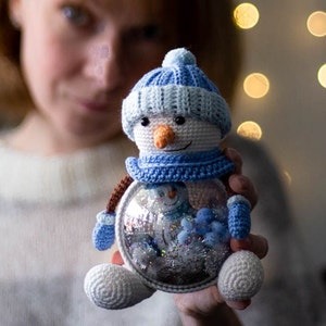 May include: A crocheted snowman ornament with a blue hat, scarf, and mittens. The snowman has an orange carrot nose and a smiling face. Inside the clear glass body is a miniature snowman and glitter. A festive winter holiday decoration.