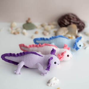 K&ouml;nnte beinhalten: Drei geh&auml;kelte Axolotl-Pl&uuml;schtiere in einer Reihe. Der Axolotl links ist lila mit einem lila Kragen, der mittlere Axolotl ist rosa mit einem rosa Kragen und der Axolotl rechts ist hellblau mit einem blauen Kragen.