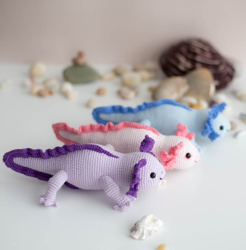 K&ouml;nnte beinhalten: Drei geh&auml;kelte Axolotl-Pl&uuml;schtiere in einer Reihe. Der Axolotl links ist lila mit einem lila Kragen, der mittlere Axolotl ist rosa mit einem rosa Kragen und der Axolotl rechts ist hellblau mit einem blauen Kragen.