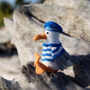 May include: A crocheted seagull toy, featuring a white body, a blue and white striped top, and a blue beret. The seagull has an orange beak and feet. The toy is resting on a piece of driftwood.