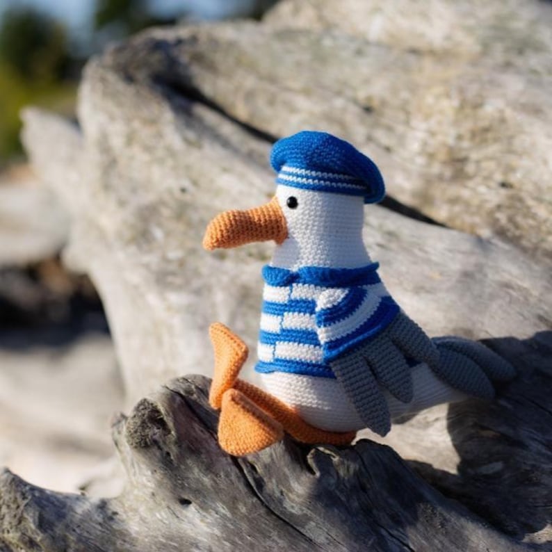 May include: A crocheted seagull toy, featuring a white body, a blue and white striped top, and a blue beret. The seagull has an orange beak and feet. The toy is resting on a piece of driftwood.