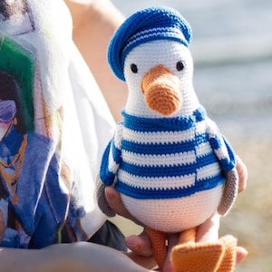 May include: A crocheted seagull toy, dressed in a blue and white striped top and a blue beret. The seagull has an orange beak and feet. The soft toy is held in someone's hands. A charming, handmade item.