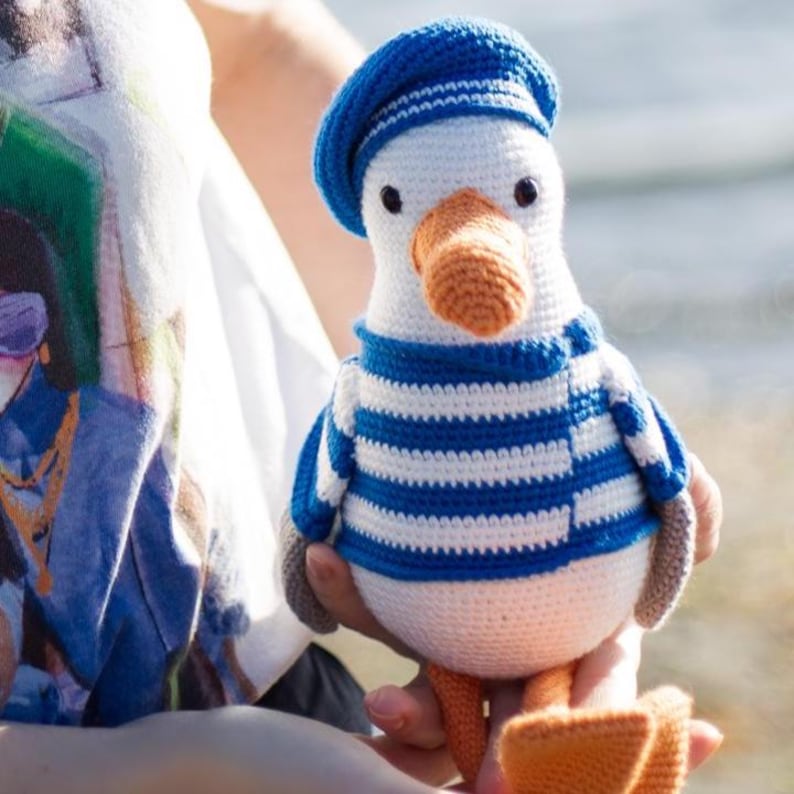 May include: A crocheted seagull toy, dressed in a blue and white striped top and a blue beret. The seagull has an orange beak and feet. The soft toy is held in someone's hands. A charming, handmade item.