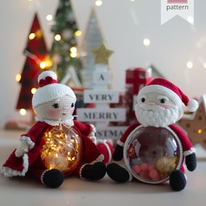 Peut inclure: Deux figurines faites &agrave; la main au crochet repr&eacute;sentant le P&egrave;re No&euml;l, avec des ventres transparents remplis de lumi&egrave;res et d'ornements. Les P&egrave;res No&euml;l portent des tenues rouges et blanches avec des bottes noires. L'arri&egrave;re-plan comprend des sapins de No&euml;l et le texte "A VERY MERRY CHRISTMAS".