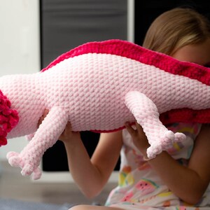 K&ouml;nnte beinhalten: Ein rosa und rotes geh&auml;keltes Axolotl-Pl&uuml;schtier. Das Spielzeug wird von einer Person gehalten.