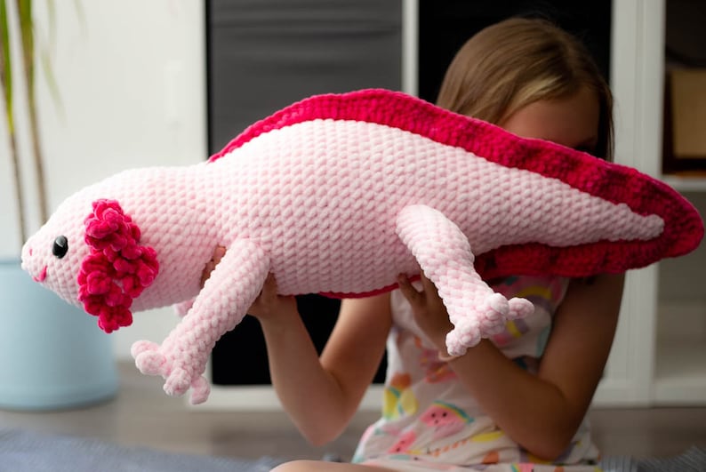 K&ouml;nnte beinhalten: Ein rosa und rotes geh&auml;keltes Axolotl-Pl&uuml;schtier. Das Spielzeug wird von einer Person gehalten.