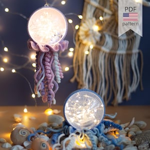 Puede incluir: Dos adornos de medusas de ganchillo hechos a mano con luces de hadas internas. Uno es morado y rosa, el otro es azul. Los adornos están sobre una mesa con conchas marinas y luces de hadas. La imagen incluye un gráfico de patrón PDF.