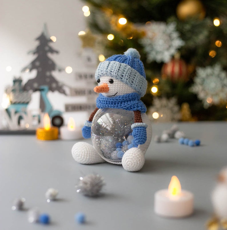 May include: A handmade crochet snowman with a blue hat, scarf, and mittens. The snowman has a carrot nose and a clear glass body filled with silver glitter and blue beads. It is a festive holiday decoration.