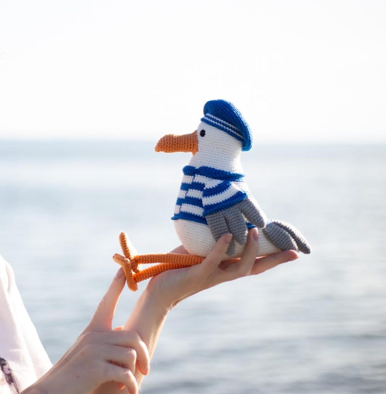 May include: A crocheted seagull toy, white with a blue and white striped top, a blue hat, grey wings, and orange beak and feet. The toy is held in a hand against a blurred sea background.