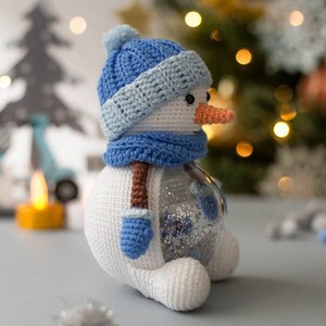 May include: A handmade crochet snowman with a blue hat, scarf, and mittens. The snowman has a carrot nose and a clear glass body filled with glitter. A festive winter holiday decoration.