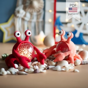 Pode incluir: Duas figuras de caranguejo em crochê, uma vermelho-brilhante e outra laranja-coral, cada uma com um corpo esférico transparente preenchido com luzes brilhantes, dois olhos elevados em hastes e duas pinças, sobre pequenas conchas marinhas.