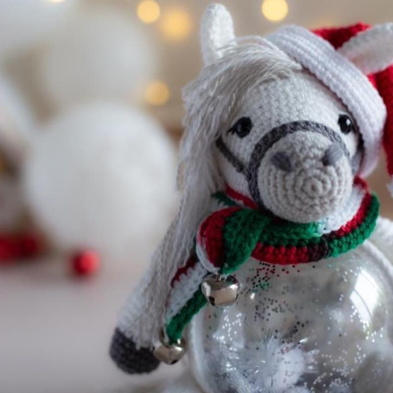 May include: A white crocheted horse ornament with a Santa hat and a scarf with red, green, and white stripes. The horse has a gray bridle and two silver bells. The ornament is inside a clear glass ball filled with glitter.