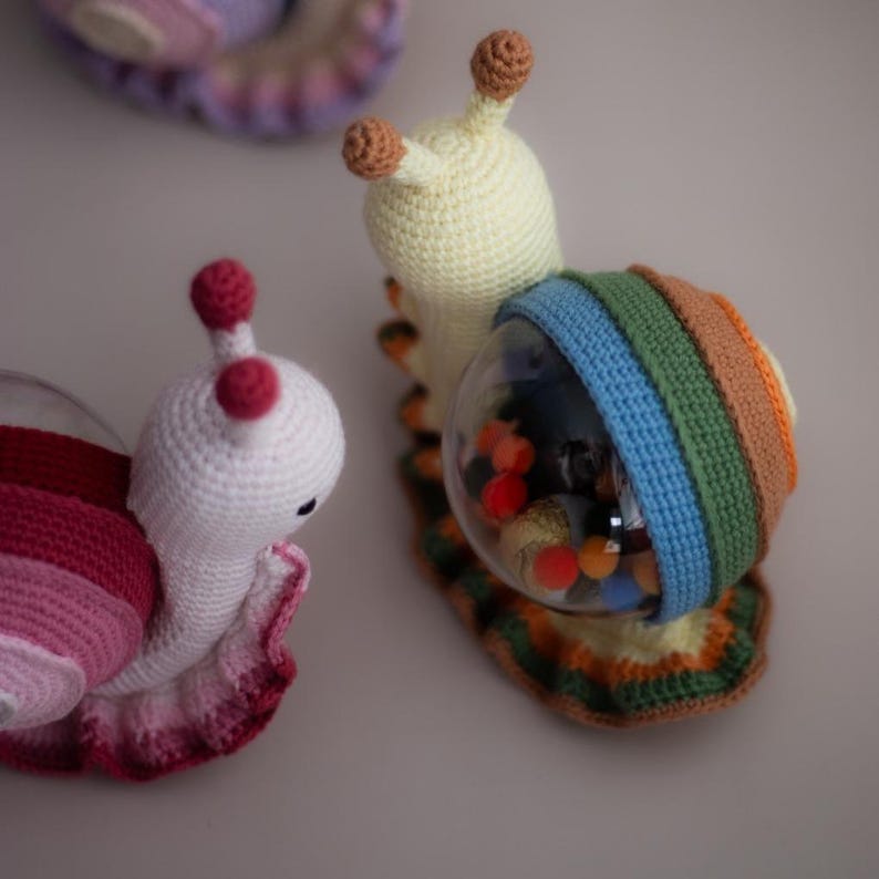 May include: Three crocheted snail toys. One is pink and white, one is yellow and white, and the third is yellow and has a clear plastic shell filled with colorful beads.