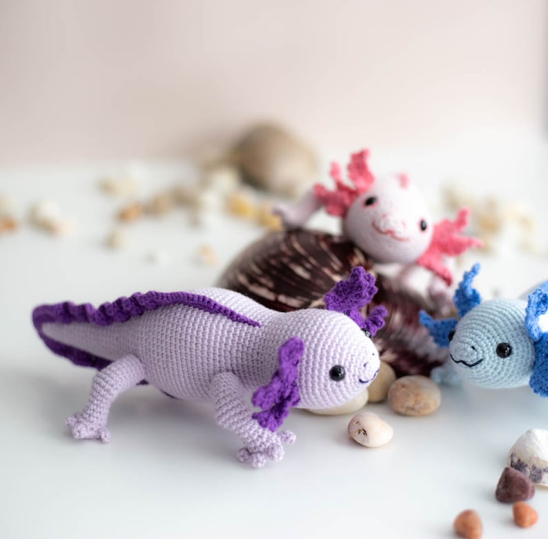 K&ouml;nnte beinhalten: Drei geh&auml;kelte Axolotl-Pl&uuml;schtiere in verschiedenen Farben. Der Axolotl links ist lila mit einem helllila K&ouml;rper, der Axolotl in der Mitte ist rosa mit einem wei&szlig;en K&ouml;rper und der Axolotl rechts ist blau mit einem hellblauen K&ouml;rper.