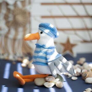 May include: A crocheted seagull toy, featuring a blue and white striped hat and sweater. The seagull has an orange beak and feet. It is resting on small stones. The toy is white, blue, grey, and orange.