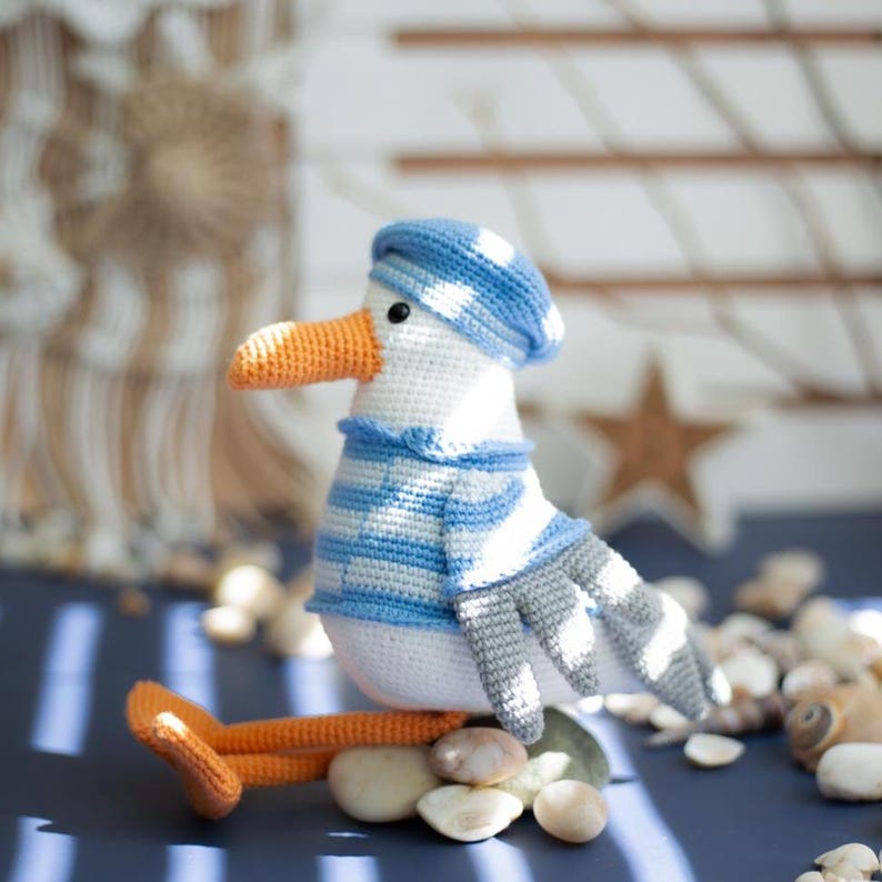 May include: A crocheted seagull toy, featuring a blue and white striped hat and sweater. The seagull has an orange beak and feet. It is resting on small stones. The toy is white, blue, grey, and orange.