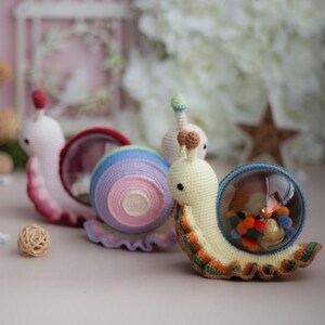 May include: Three crocheted snail toys. The first snail is pink with a red shell. The second snail is purple, blue, and pink with a rainbow shell. The third snail is yellow with a green shell and a clear plastic dome containing a small crocheted orange and brown animal.