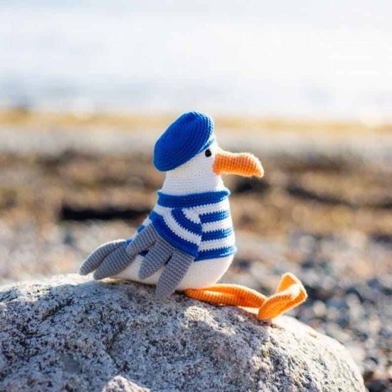 May include: A handcrafted crochet seagull toy, sporting a blue beret and a blue and white striped sweater. The seagull has grey wings, an orange beak and feet, and is perched on a grey rock. The background is blurred.