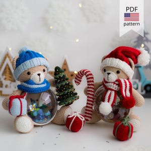 Op de afbeelding: Twee gehaakte teddyberen gekleed in winterkleding. Eén beer draagt ​​een blauwe muts en sjaal, terwijl de andere een rode kerstmanmuts en sjaal draagt. Beide beren houden een heldere glazen balversiering vast met glitter en andere decoraties erin. De beren zitten op een wit oppervlak met een kleine kerstboom, een snoepstok en een rode geschenkdoos.