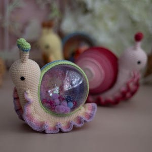 May include: A crocheted snail toy with a clear plastic shell filled with colorful pom-poms and glitter. The snail is pink, purple, and green.