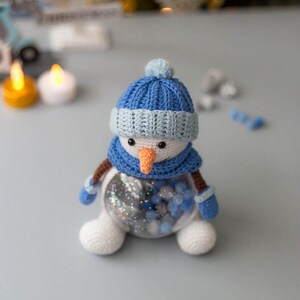 May include: A handmade crochet snowman with a blue hat, scarf, and mittens. The snowman has a carrot nose and black eyes. The clear body is filled with blue and silver decorative beads. A charming winter holiday decoration.