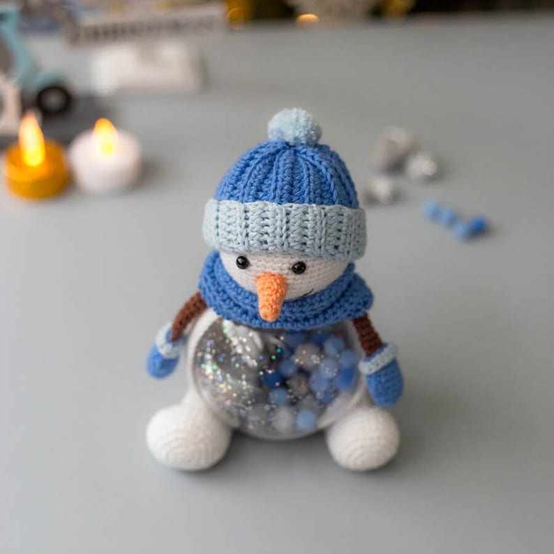May include: A handmade crochet snowman with a blue hat, scarf, and mittens. The snowman has a carrot nose and black eyes. The clear body is filled with blue and silver decorative beads. A charming winter holiday decoration.