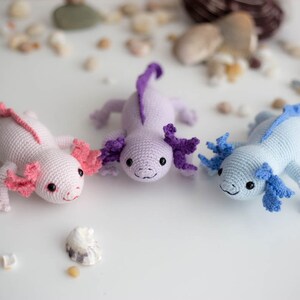 K&ouml;nnte beinhalten: Drei geh&auml;kelte Axolotl-Pl&uuml;schtiere in Rosa, Lila und Blau. Die Axolotl l&auml;cheln alle und haben gro&szlig;e, runde Augen.