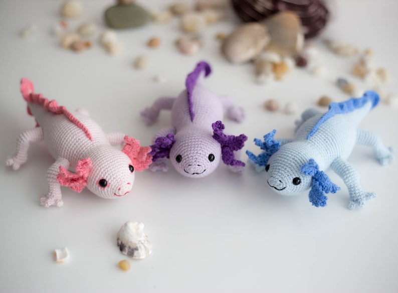 K&ouml;nnte beinhalten: Drei geh&auml;kelte Axolotl-Pl&uuml;schtiere in Rosa, Lila und Blau. Die Axolotl l&auml;cheln alle und haben gro&szlig;e, runde Augen.