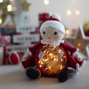 Peut inclure: Poup&eacute;e au crochet faite &agrave; la main, v&ecirc;tue d'un costume de P&egrave;re No&euml;l rouge et blanc. Le corps de la poup&eacute;e est une boule en verre transparente remplie de guirlandes lumineuses blanches chaudes. Elle a un chapeau et un col blancs, et des chaussures noires.