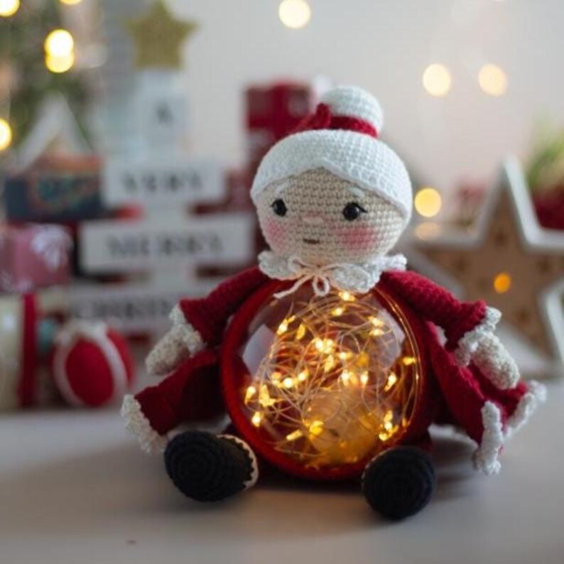 Peut inclure: Poup&eacute;e au crochet faite &agrave; la main, v&ecirc;tue d'un costume de P&egrave;re No&euml;l rouge et blanc. Le corps de la poup&eacute;e est une boule en verre transparente remplie de guirlandes lumineuses blanches chaudes. Elle a un chapeau et un col blancs, et des chaussures noires.