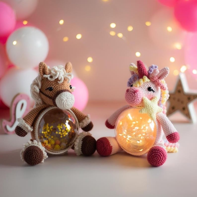 May include: Two crocheted plush toys: a brown horse and a pink unicorn. Each has a clear, round belly filled with confetti and a warm light. The unicorn holds a star. Balloons and fairy lights create a festive background.