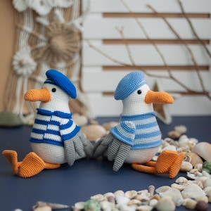 May include: Two crocheted seagull dolls, each wearing a blue beret and a blue and white striped top. They have orange beaks and feet, grey wings, and black eyes. The dolls are on a blue surface, surrounded by seashells.