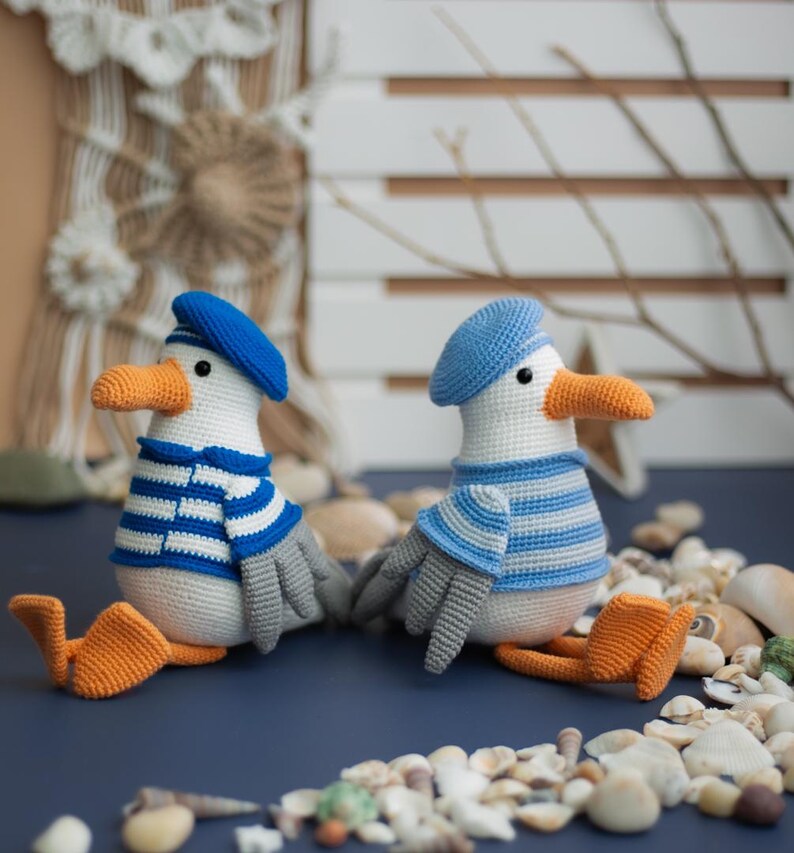 May include: Two crocheted seagull dolls, each wearing a blue beret and a blue and white striped top. They have orange beaks and feet, grey wings, and black eyes. The dolls are on a blue surface, surrounded by seashells.