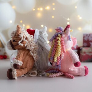 May include: Three crocheted plush toys: a brown horse, a white horse with a red hat, and a pink unicorn. The horse toys have tan manes and tails. The unicorn has a rainbow mane and tail. All three are sitting on a white surface.
