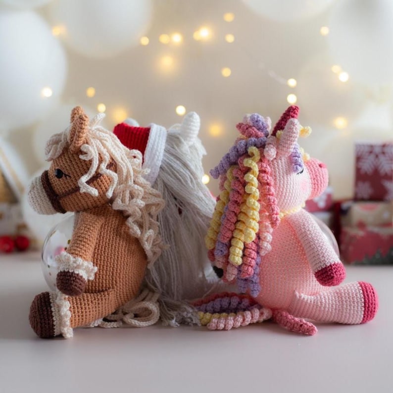 May include: Three crocheted plush toys: a brown horse, a white horse with a red hat, and a pink unicorn. The horse toys have tan manes and tails. The unicorn has a rainbow mane and tail. All three are sitting on a white surface.