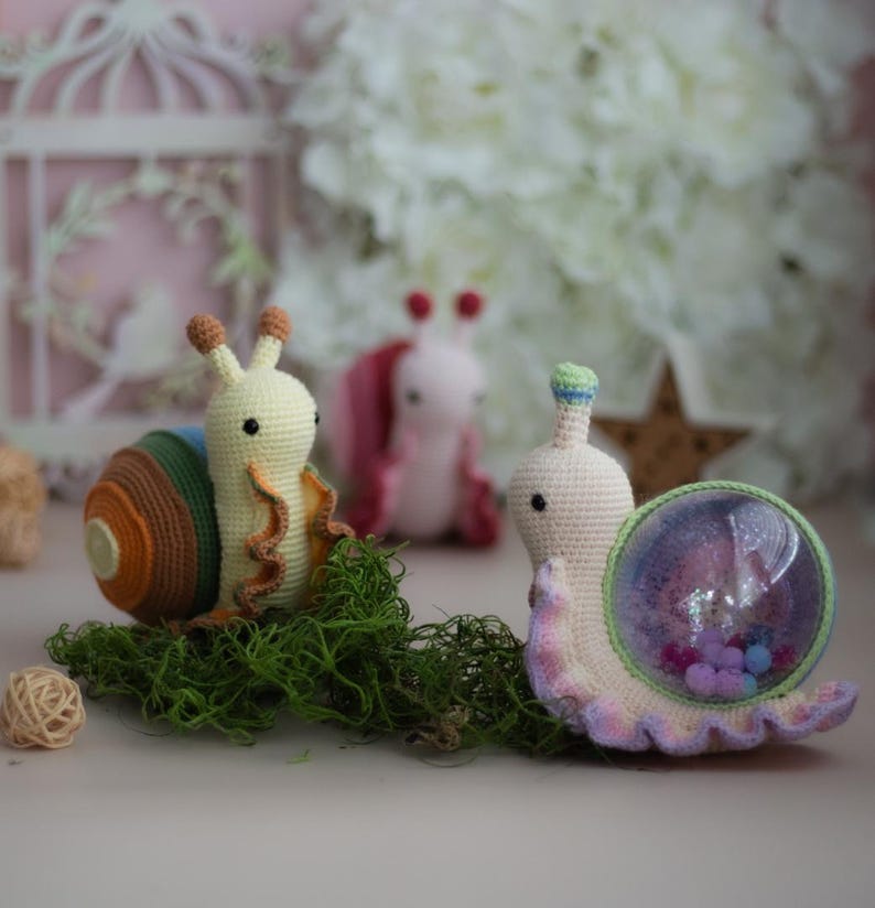 May include: Three crocheted snail toys. One snail is yellow with brown stripes, one is pink with green accents, and one is white with purple and green accents. The white snail has a clear plastic shell filled with colorful beads.