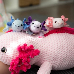 K&ouml;nnte beinhalten: Ein rosa geh&auml;keltes Axolotl-Pl&uuml;schtier mit drei kleineren geh&auml;kelten Axolotl-Pl&uuml;schtieren auf dem R&uuml;cken. Die kleineren Pl&uuml;schtiere sind blau, lila und rosa.