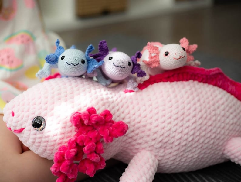 K&ouml;nnte beinhalten: Ein rosa geh&auml;keltes Axolotl-Pl&uuml;schtier mit drei kleineren geh&auml;kelten Axolotl-Pl&uuml;schtieren auf dem R&uuml;cken. Die kleineren Pl&uuml;schtiere sind blau, lila und rosa.